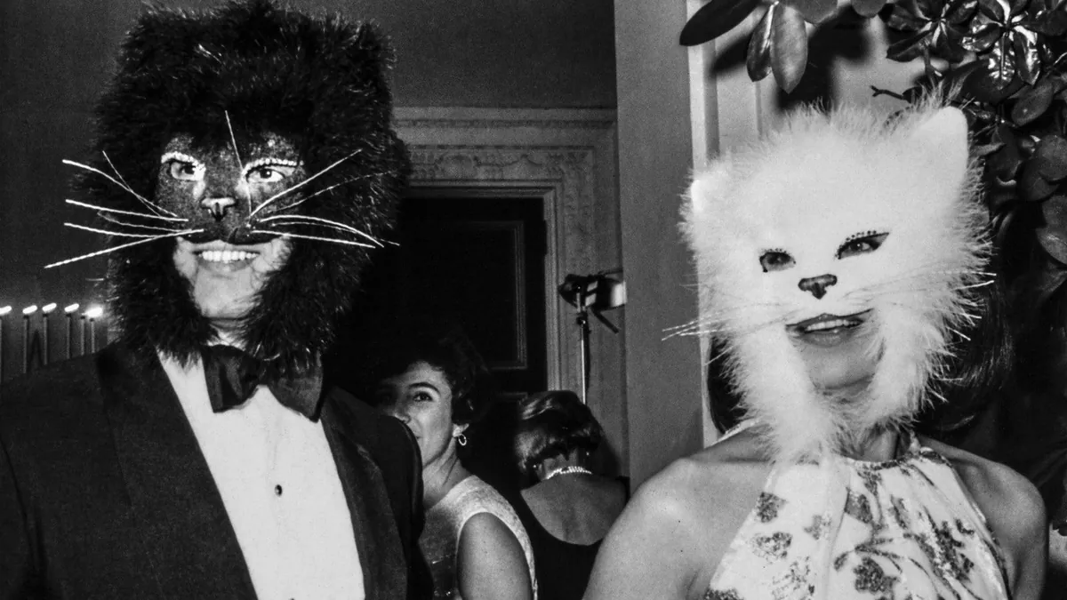 Two guests in furred cat masks at a black-tie ball, caught by hard on-camera flash in black and white