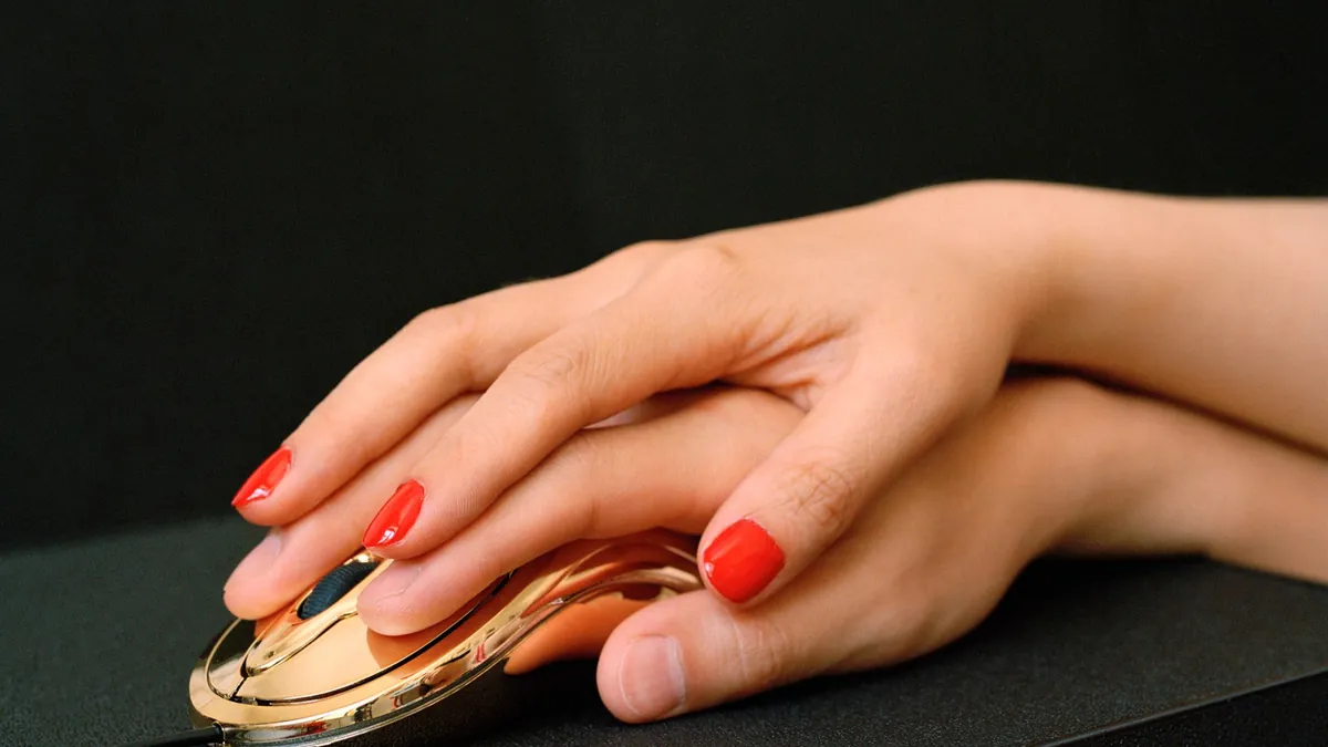 A hand with red nails presses down on a gold mouse against a dark desk, lit hard and close