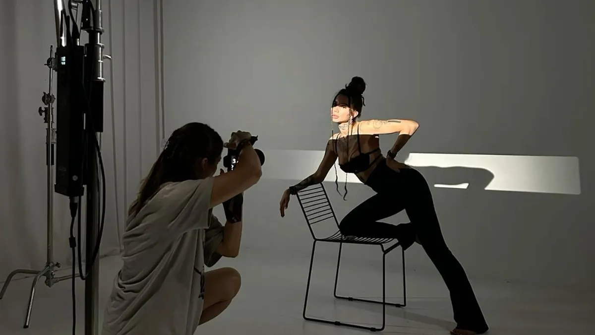 A photographer kneels to shoot a model on a grey cyclorama, light stand and flag standing in frame