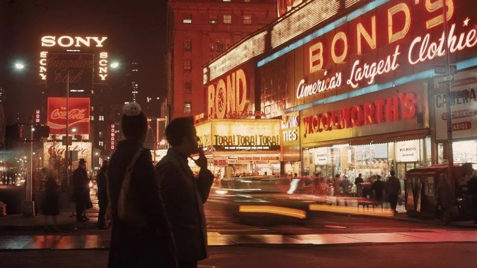 Two figures wait on a neon-lit street corner at night as a taxi streaks past in a long exposure