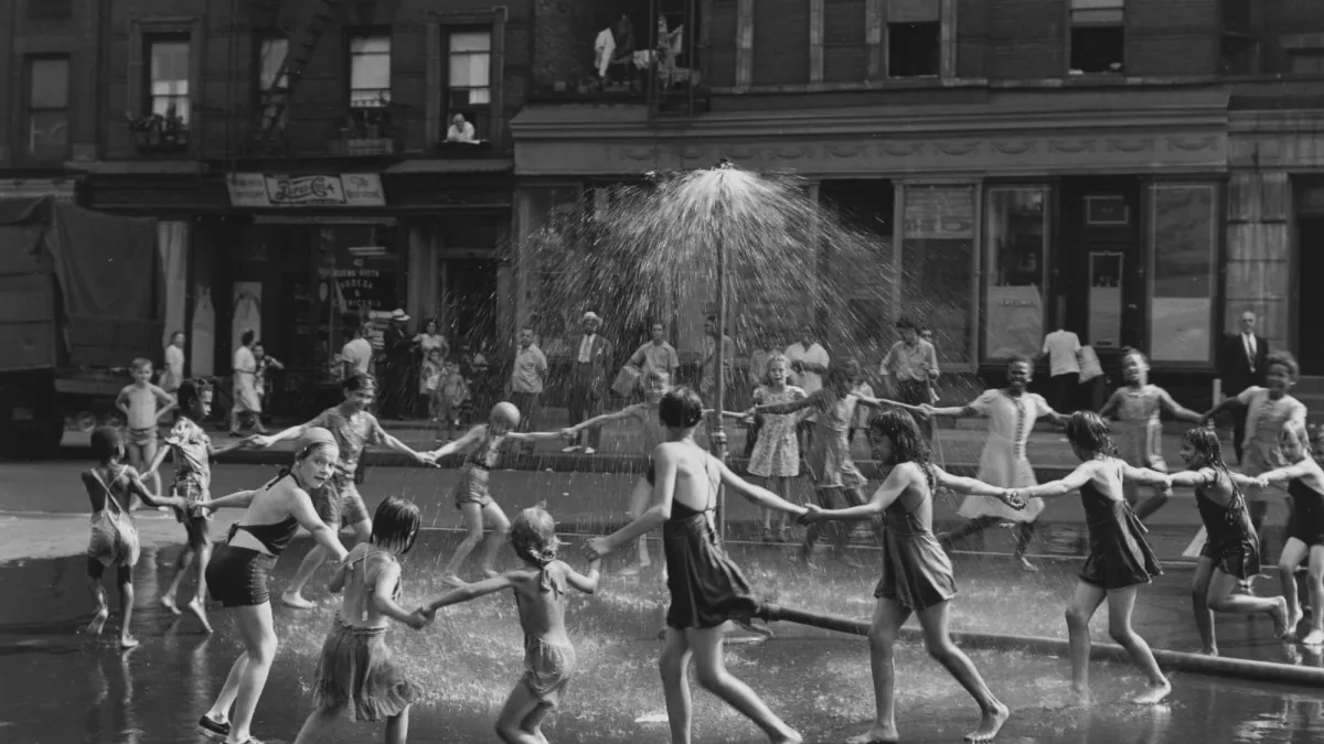 Children run through a street hydrant's spray in front of tenement blocks, mid-stride, in black and white