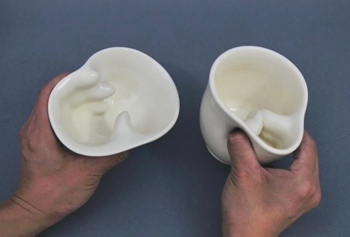 Two hands lifting a pair of white ceramic vessels from a grey studio surface