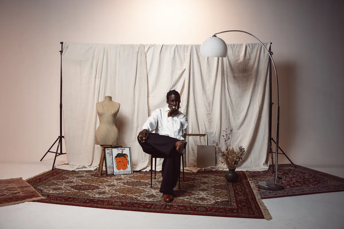 A person seated in front of a draped backdrop and lamp on a patterned rug, a small home set