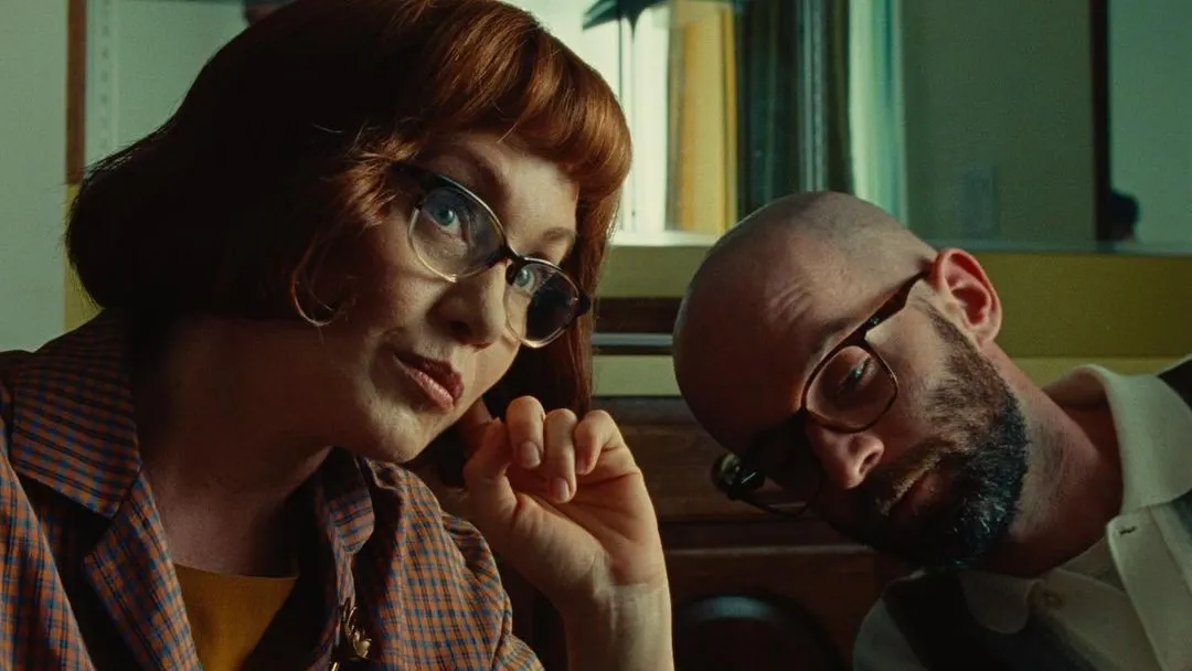 Two actors close together in a warm interior, a red-haired woman in glasses beside a man mid-scene